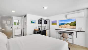 A modern hotel room with a bed, a desk, a chair, a small kitchenette, and a large window with a beach view.