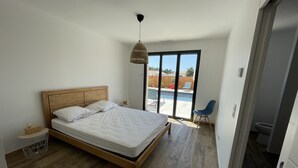 A bedroom with a wooden bed, a nightstand, a hanging light, and a view of a swimming pool through a glass door. - Seaside villa with large pool and terrace (Vendays-Montalivet)