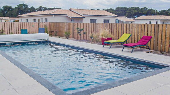 A rectangular swimming pool with a tiled deck, surrounded by a wooden fence and a row of colorful lounge chairs.