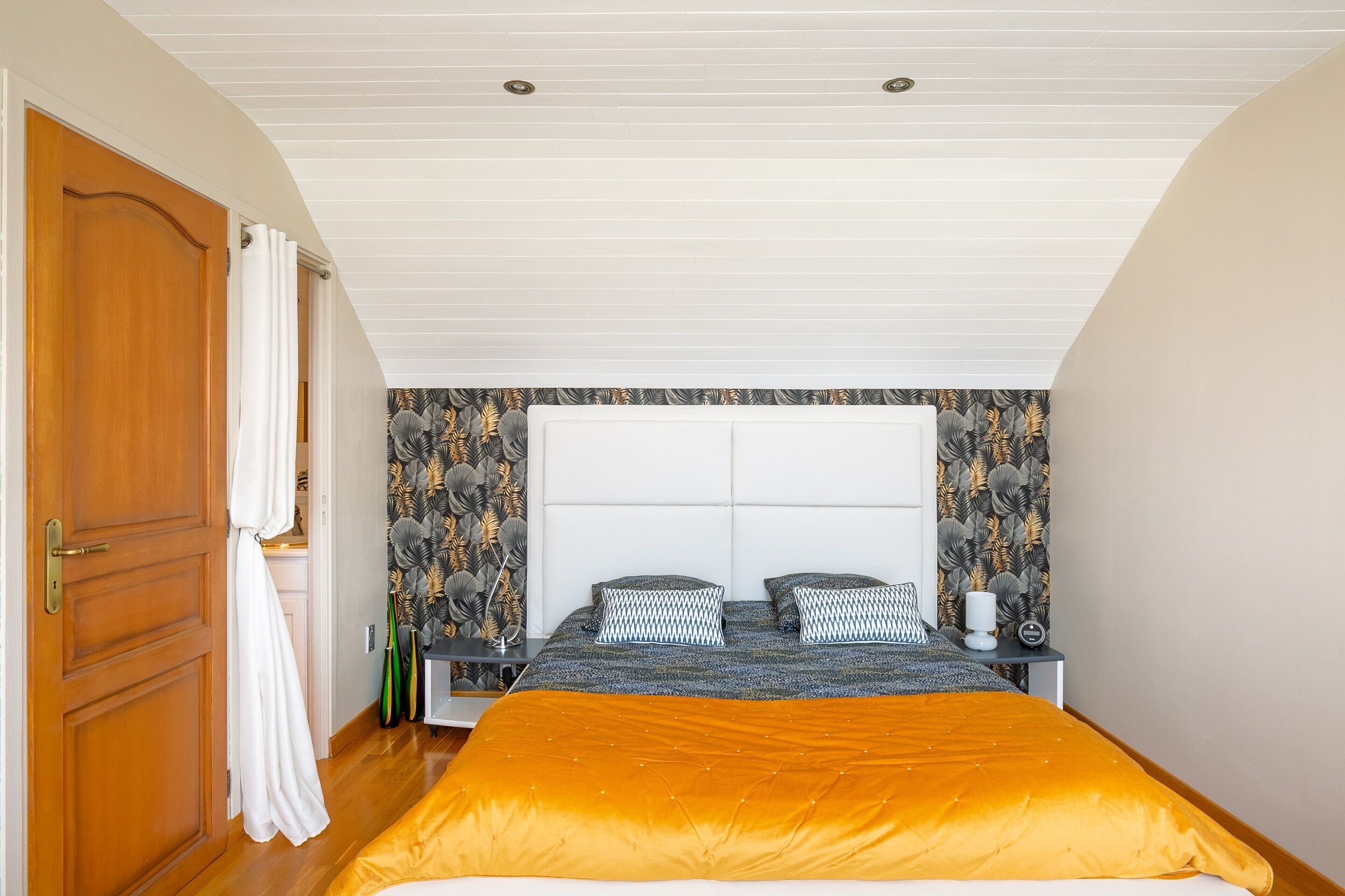A bedroom with a bed, a nightstand, a wall-mounted headboard, and a door.