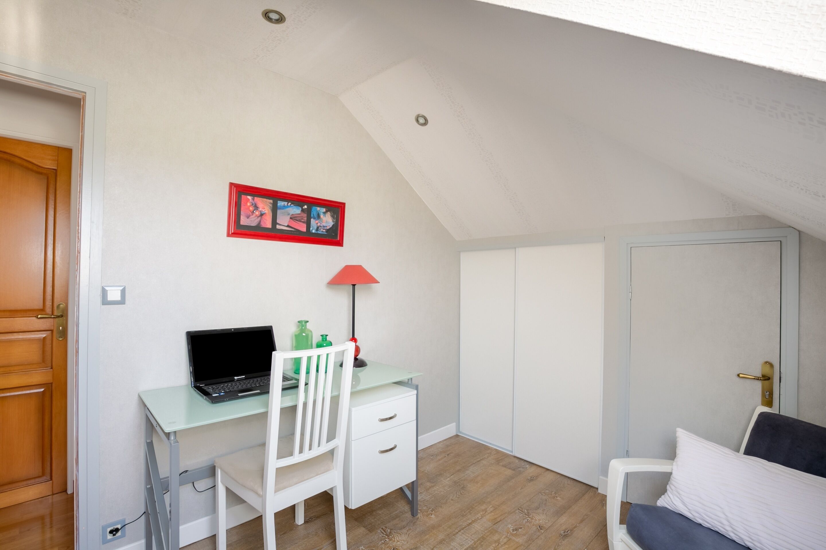 A small, well-lit room with a desk, chair, and a bed.