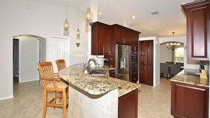 A kitchen with dark wood cabinets, a granite countertop, and stainless steel appliances. There are two wooden chairs and a bar stool. - Newly Furnished, Solar Heated pool, Game room, 10 mins from Beach (Sebastian)