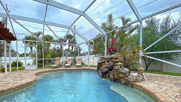A swimming pool with a glass enclosure, surrounded by lounge chairs and a stone waterfall feature. - Newly Furnished, Solar Heated pool, Game room, 10 mins from Beach (Sebastian)