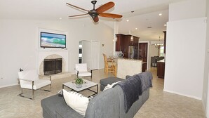 A living room with a gray sofa, a white armchair, a coffee table, and a fireplace. There is a ceiling fan, a kitchen in the background, and a dining area with wooden chairs. - Newly Furnished, Solar Heated pool, Game room, 10 mins from Beach (Sebastian)