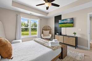 A bedroom with a large bed, a ceiling fan, a television, a wicker chair, and a view of greenery through the window.
