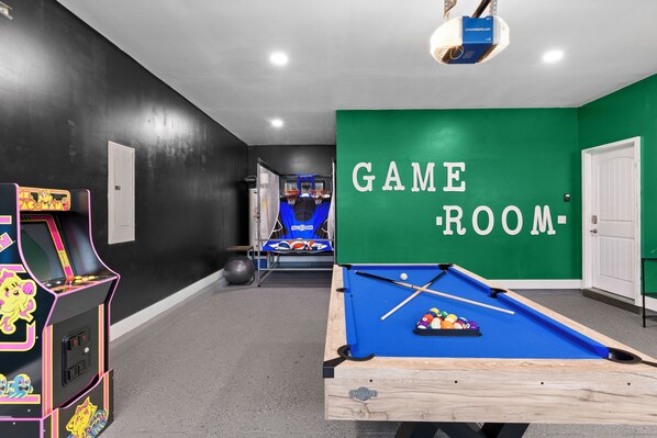 A game room with a pool table, a pool cue, and a pool table with balls. There is a neon arcade machine and a basketball hoop in the background.