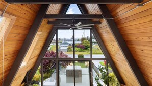 A high-ceilinged room with a sloped wooden roof, a large window offering a view of the outdoors, and a ceiling fan.