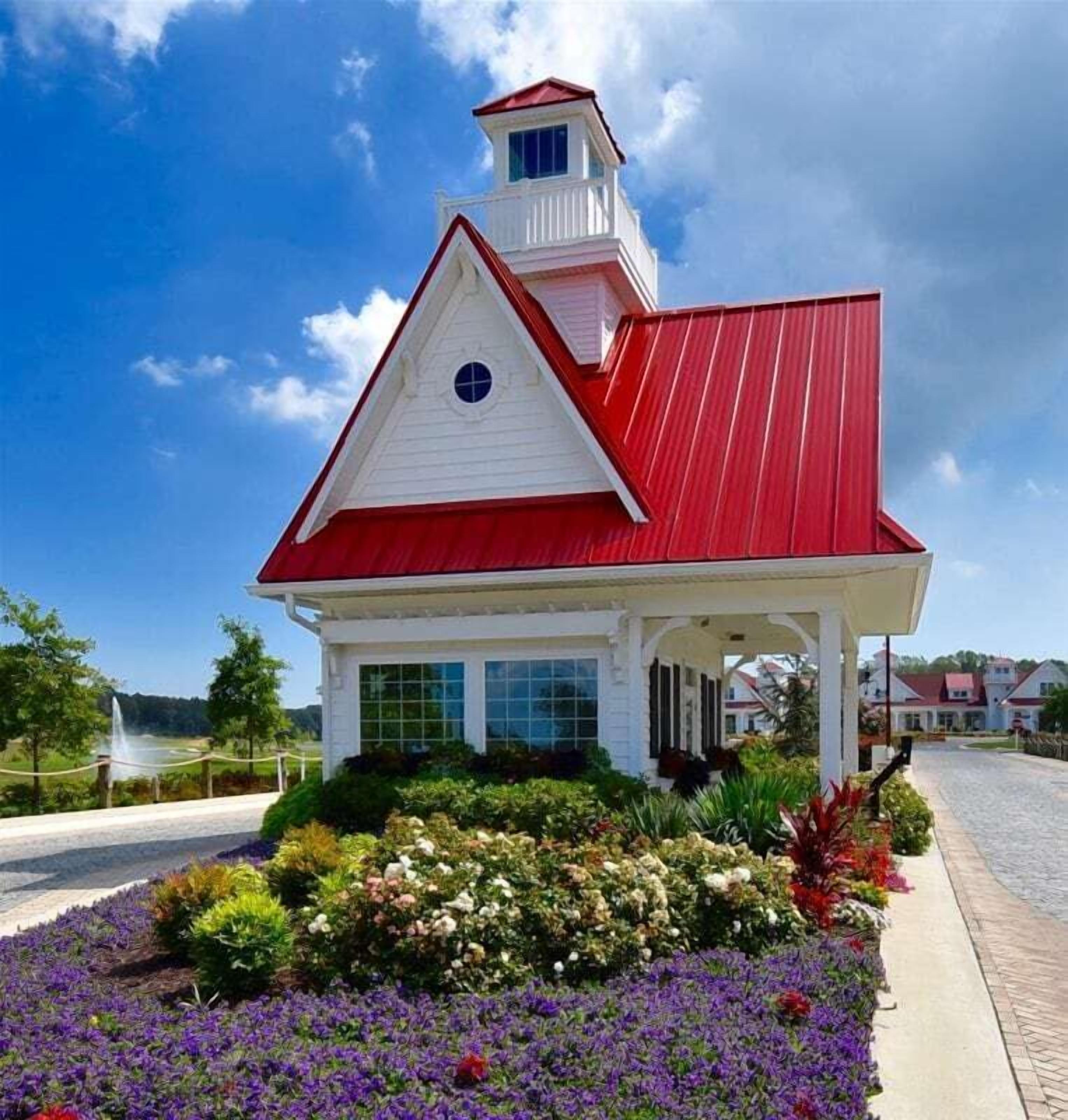 Un bâtiment blanc avec un toit rouge et une petite tour, entouré d’un jardin où poussent différentes sortes de fleurs.