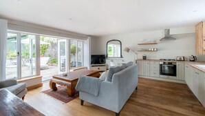 Un vaste espace de vie avec un canapé gris, une table basse en bois et un téléviseur en arrière-plan. On aperçoit une cuisine avec des meubles blancs et une cuisinière.