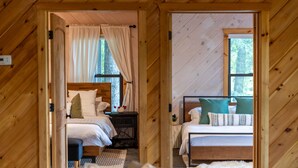 A wooden cabin bedroom with two beds, a nightstand, and a window with curtains. - Luxury 5 BR Cabin with Hot Tub and Views!  (Townsend)