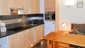 Una cucina compatta con mobili in legno, un piano di lavoro nero e un piccolo frigorifero. In un'area soggiorno adiacente si trova un tavolo da pranzo con una lampada e un divano.