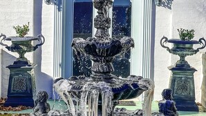 A fountain with water cascading down, surrounded by stone planters and two decorative urns, in front of a building with a white facade and a window with a decorative grille. - Smithmore Castle (Spruce Pine)
