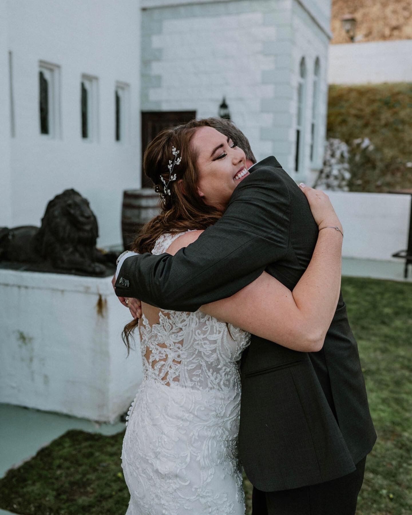 A couple embracing in front of a white building with a stone facade and a statue in the background.