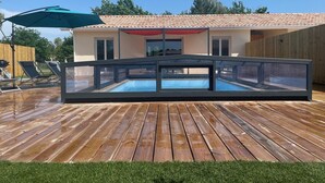 Une piscine dotée d’une structure en verre, d’une terrasse en bois et, au fond, d’un bâtiment avec un toit recouvert de tuiles.