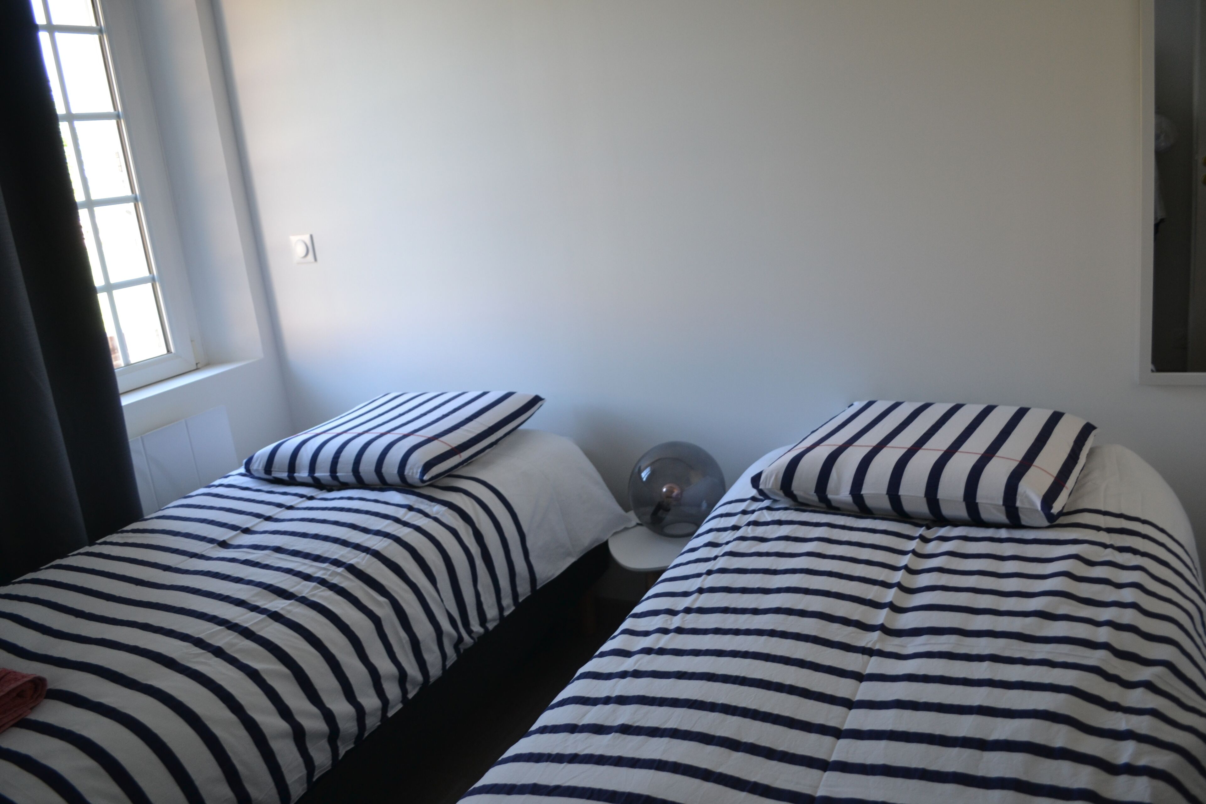 Et soveværelse med to senge, begge med stribet sengetøj, et natbord med lampe og et vindue med gardiner.