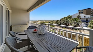 Een balkon met een houten tafel en stoelen, een decoratieve glazen vaas en uitzicht op het strand en gebouwen.