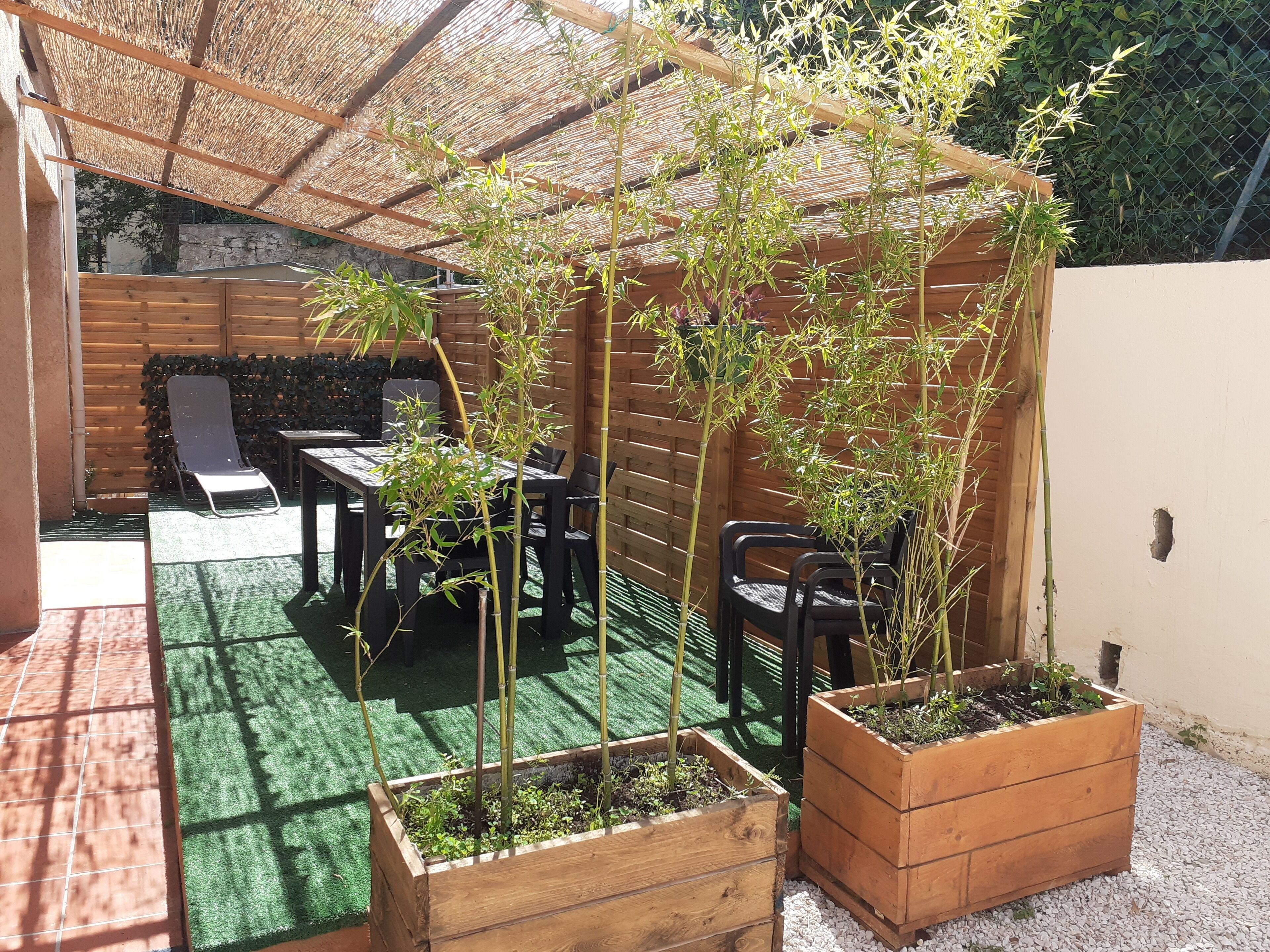 A patio area with a wooden table and chairs, a thatched roof, and bamboo plants in wooden planters.
