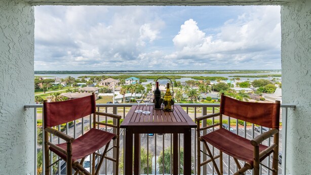 A balcony with a table and two chairs, overlooking a scenic view of a coastal town with houses and greenery. - 3 min walk to beach! Ocean and river views. Great sunsets! Updated 2 br 2ba (Daytona Beach Shores)
