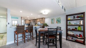 A dining area with a table and chairs, a kitchen in the background, and a bookshelf with various items. - 3 min walk to beach! Ocean and river views. Great sunsets! Updated 2 br 2ba (Daytona Beach Shores)