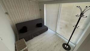 Ein Zimmer mit grauem Sofa, einem Korbstuhl und einem Kleiderständer in der Nähe eines Fensters mit Jalousien.