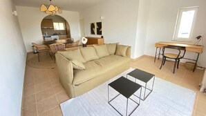 A living room with a beige sofa, two black coffee tables, a dining area with wicker chairs, a kitchen in the background, and a large window with a view. - Serenity & design T3, 2 terraces seaview 600m beach (Praia da Luz)