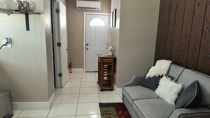 A living room with a grey sofa, a small wooden side table, a white door, and a tiled floor. - Green Castle Suite in Tampa (Tampa)