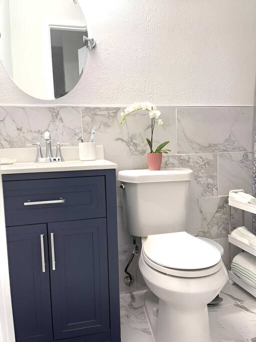 Un bagno con un water bianco, un mobile portaoggetti blu scuro, piastrelle in marmo e un'orchidea in vaso.