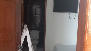 A small room with a wooden door, a toilet, a bed with a blue blanket, and a TV on the wall. - POUSADA MORADAS (Aracaju)