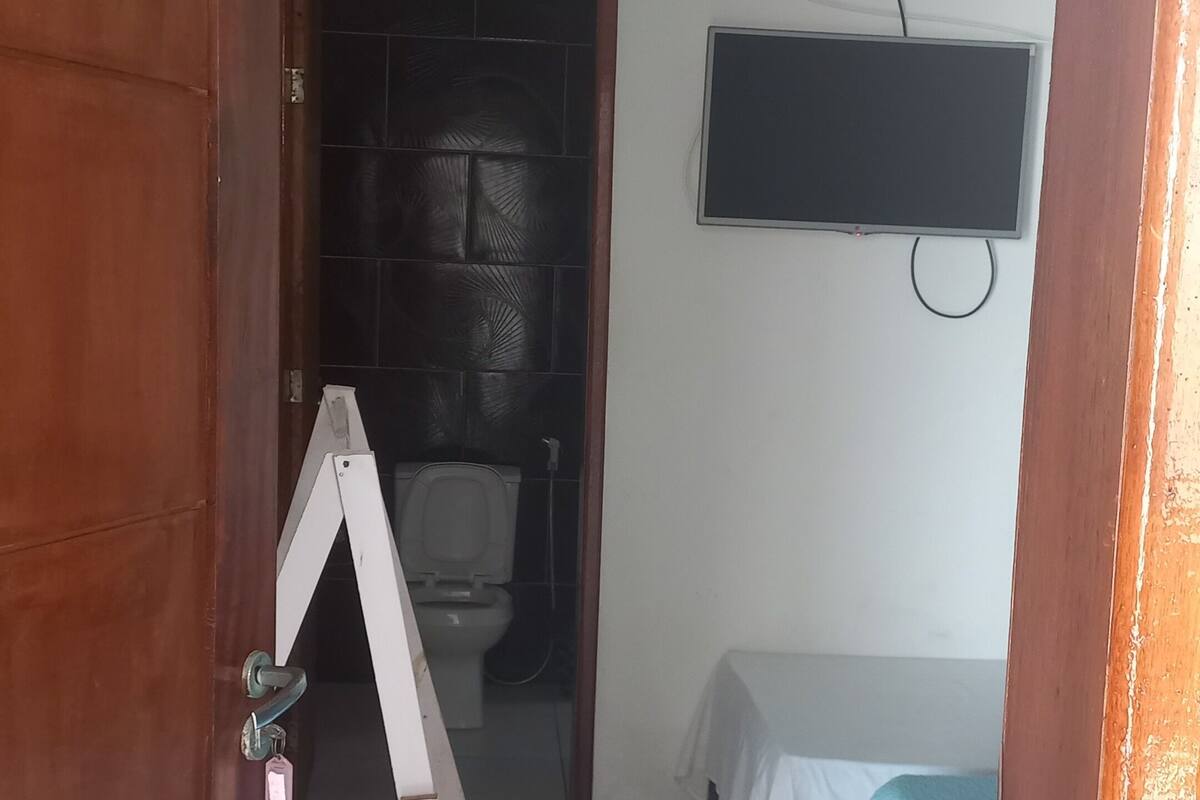 A small room with a wooden door, a toilet, a bed with a blue blanket, and a TV on the wall.