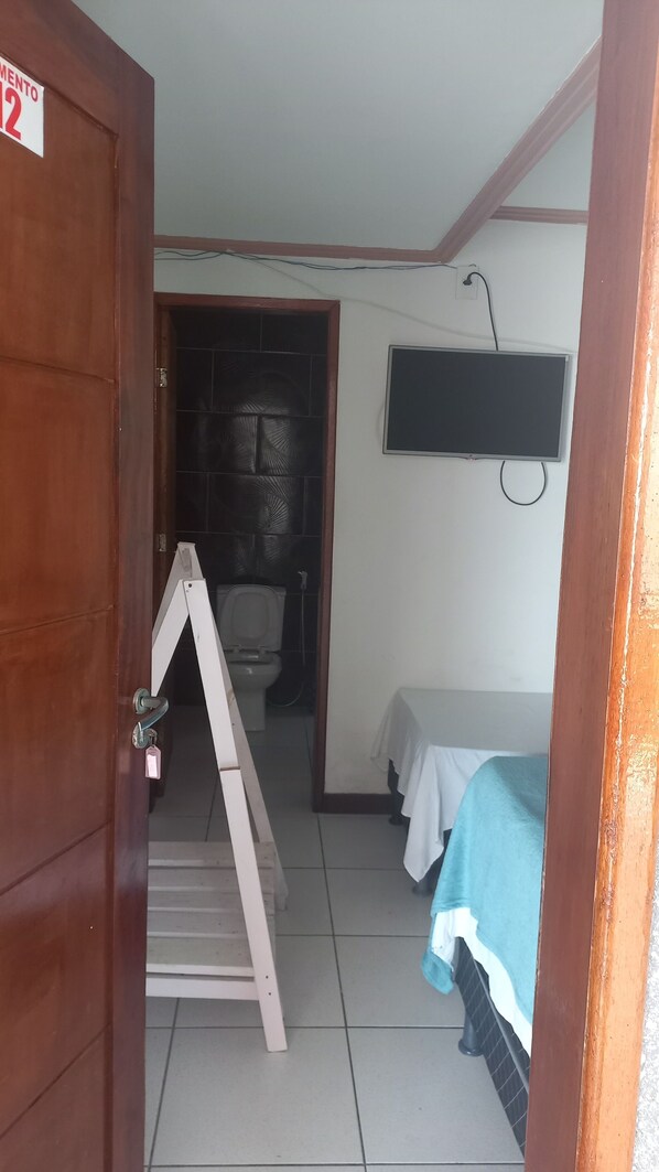Una habitación pequeña con puerta de madera, un inodoro, una cama con manta azul y un televisor en la pared.