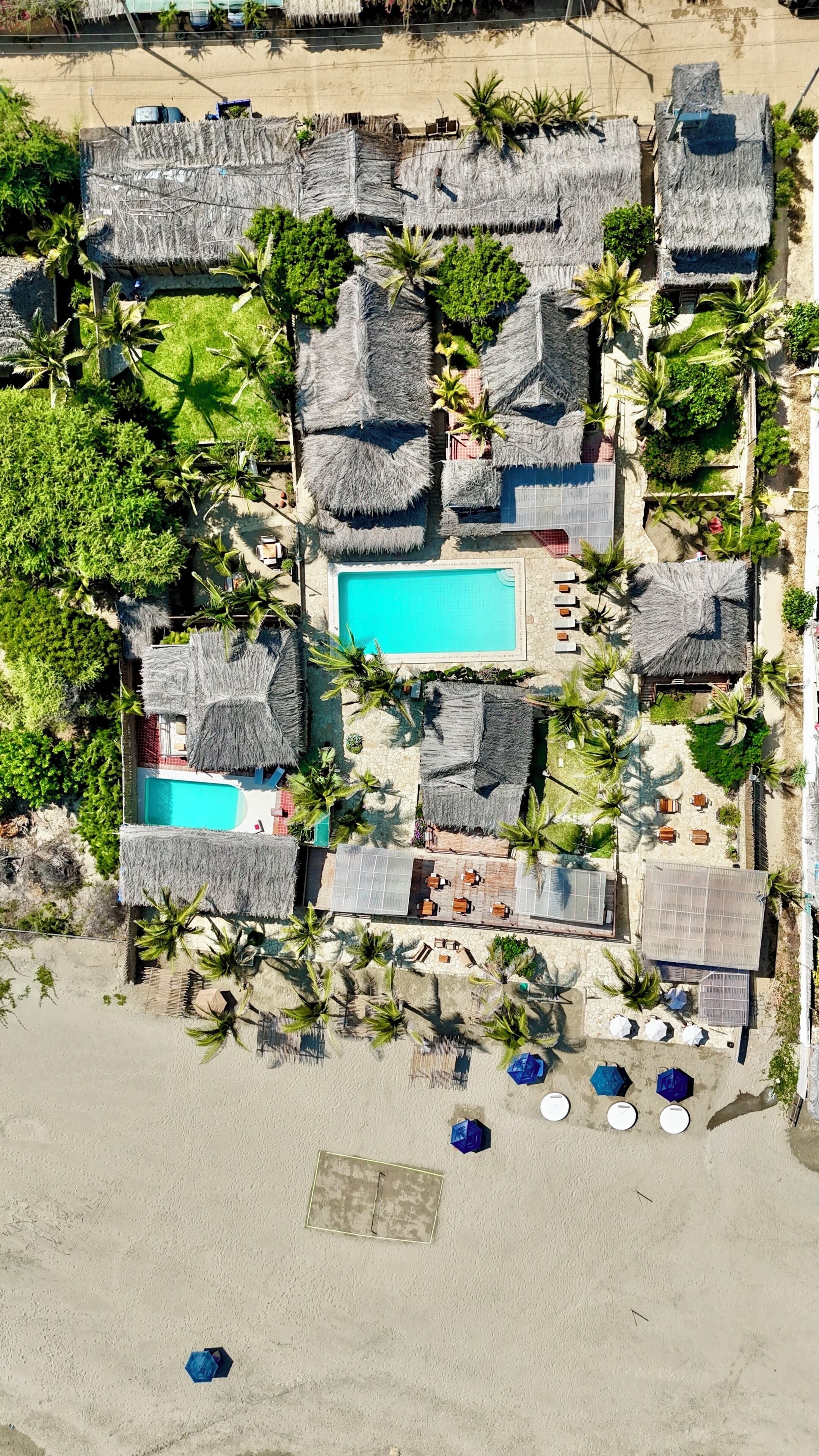 Vista aérea de um resort com chalés de telhado de palha, piscina central e área de praia arenosa com guarda-sóis.
