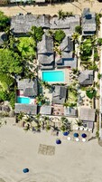 Vista aérea de un resort con cabañas de techo de paja, una piscina central y una zona de playa arenosa con sombrillas.