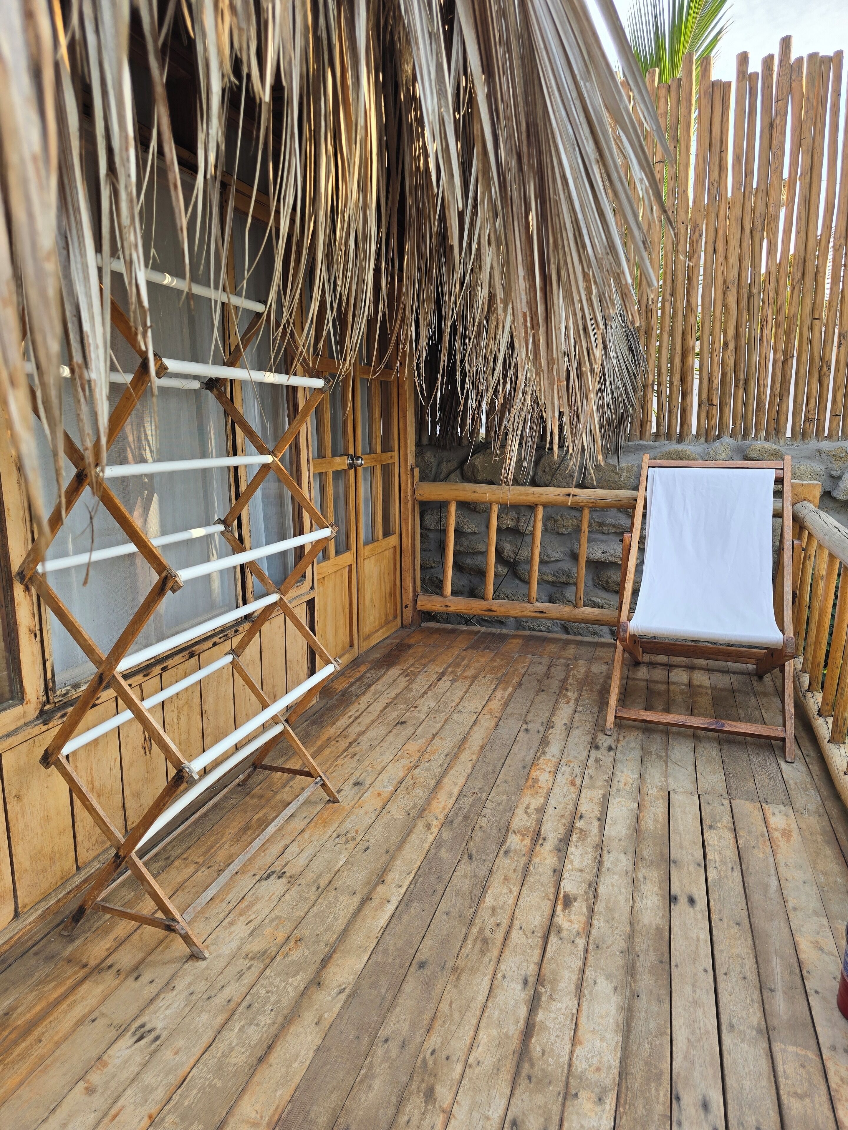 A wooden deck with a drying rack, a wooden chair with a white cushion, and a thatched roof structure.