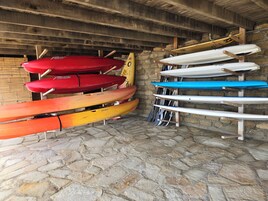 A row of kayaks and paddleboards leaning against a stone wall in a covered area.