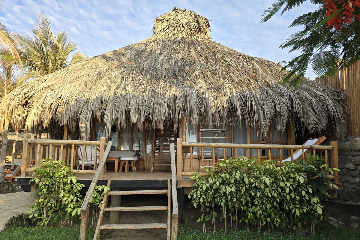 Une cabane à toit de chaume dotée d’une terrasse en bois, d’un escalier et d’un petit jardin.