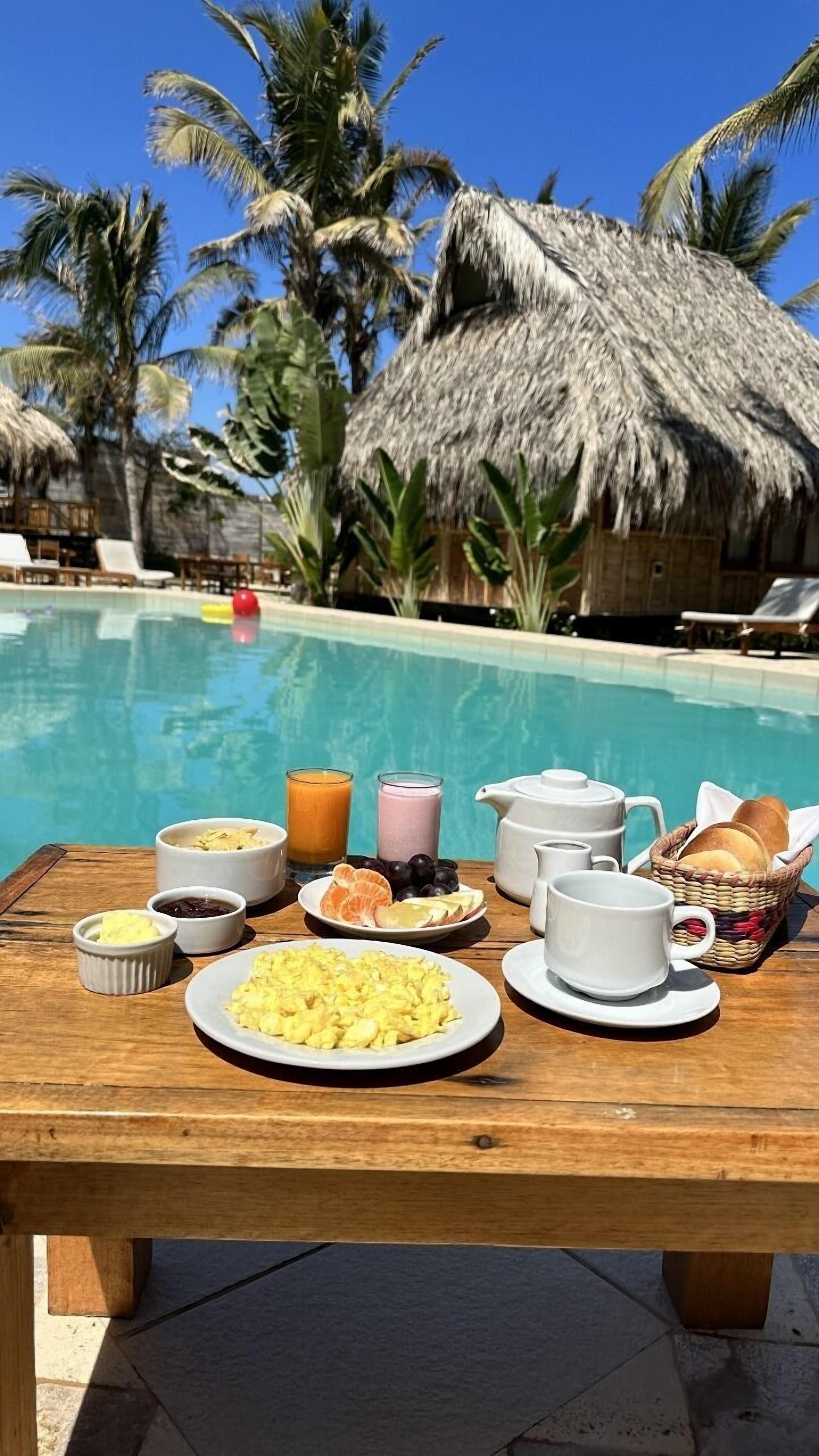 A breakfast table with eggs, toast, fruit, and beverages by a pool with palm trees and a thatched-roof hut in the background.