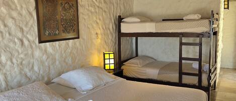 Quarto com parede branca texturizada, cama com roupa de cama branca e toalhas enroladas, beliche e luminária fixa na parede.