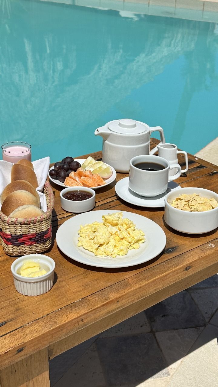 Un desayuno junto a la piscina, con pan, huevos, frutas, café y una tetera.