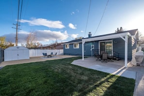 A backyard with a covered patio, a white fence, and a lawn.
