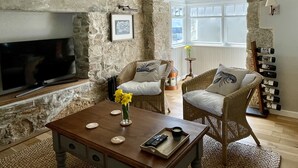 A cozy living room with stone walls, a flat-screen TV, wicker furniture, and a coffee table with a vase of yellow flowers. - Cosy & elegant cottage in charming Newlyn (Penzance)