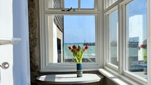 A window with a view of the sea and a lighthouse, a white door, and a small table with a vase of tulips. - Cosy & elegant cottage in charming Newlyn (Penzance)