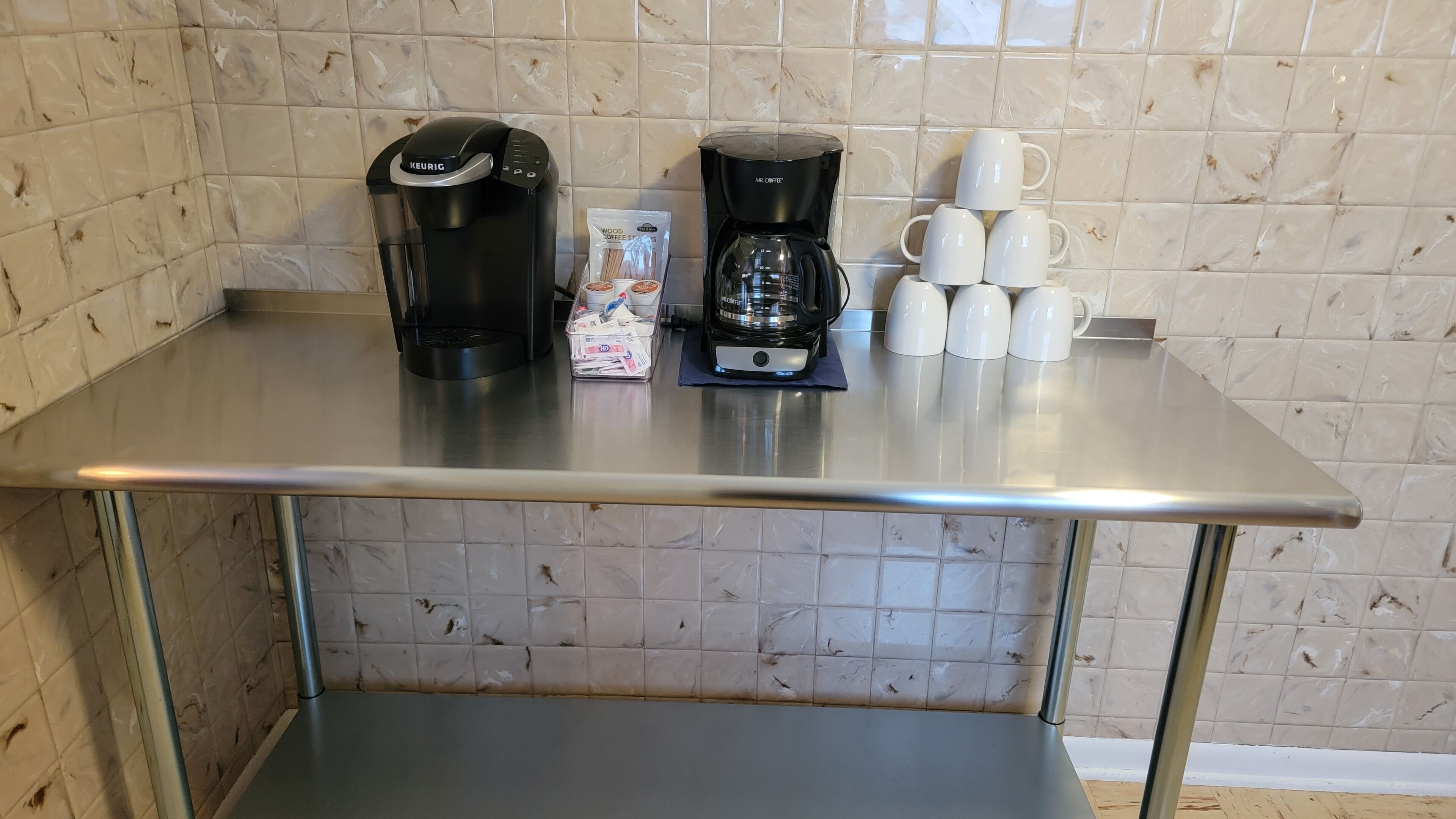 Un comptoir de cuisine avec deux machines à café, un paquet de capsules de café et plusieurs tasses blanches.