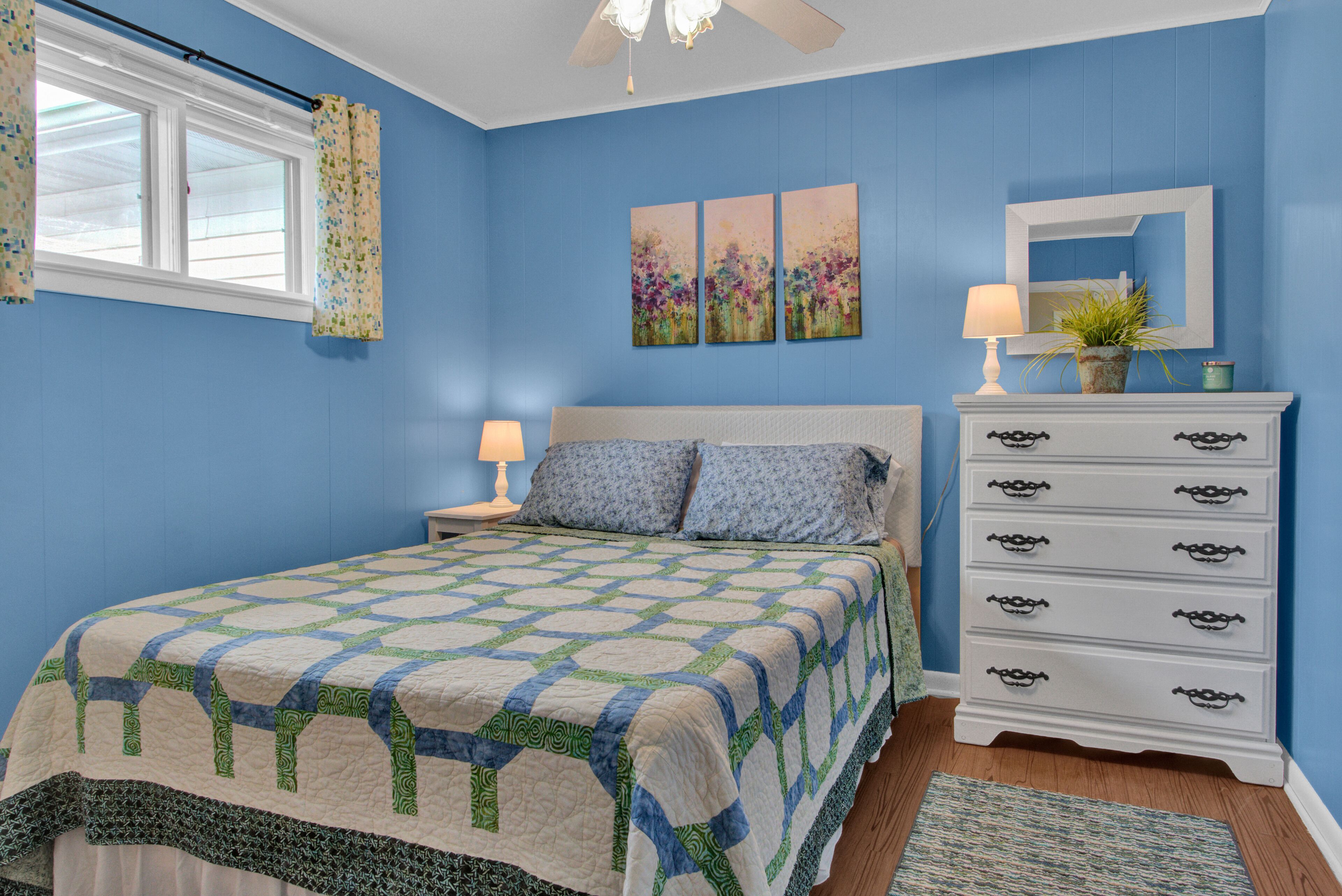 A bedroom with a bed, a dresser, a nightstand, a lamp, and a window with curtains.
