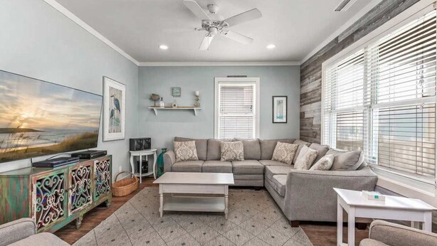 A living room with a sectional sofa, a flat-screen TV, a ceiling fan, and a window with blinds.