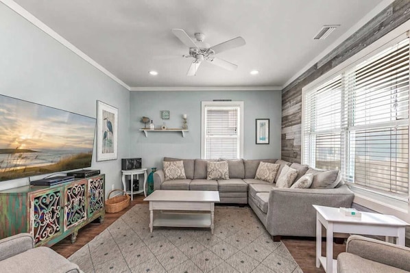 A living room with a sectional sofa, a flat-screen TV, a ceiling fan, and a window with blinds.