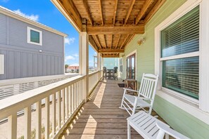 A covered wooden deck with white railings, a seating area with chairs, and a view of neighboring buildings.