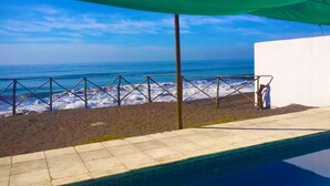Vista a la playa con barandal metálico, piscina y un edificio blanco.