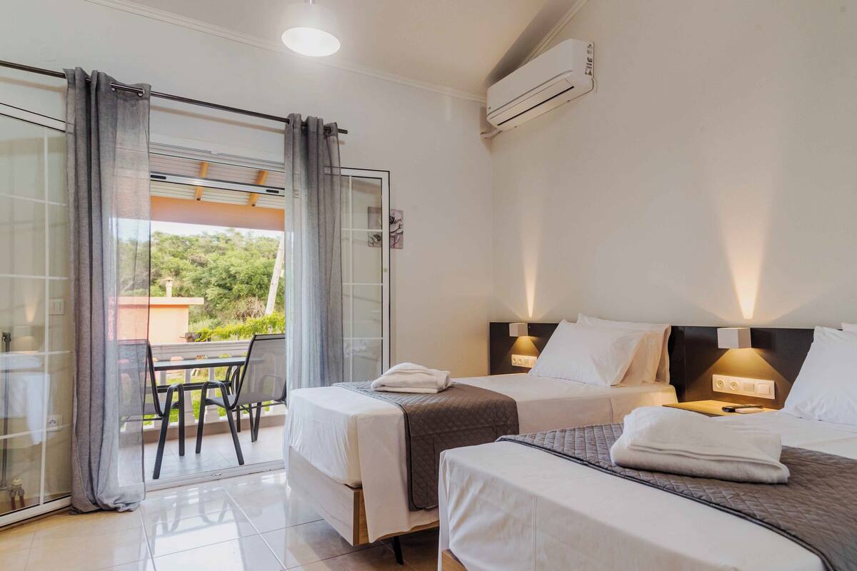 A hotel room with two beds, a balcony with a table and chairs, and an air conditioning unit.