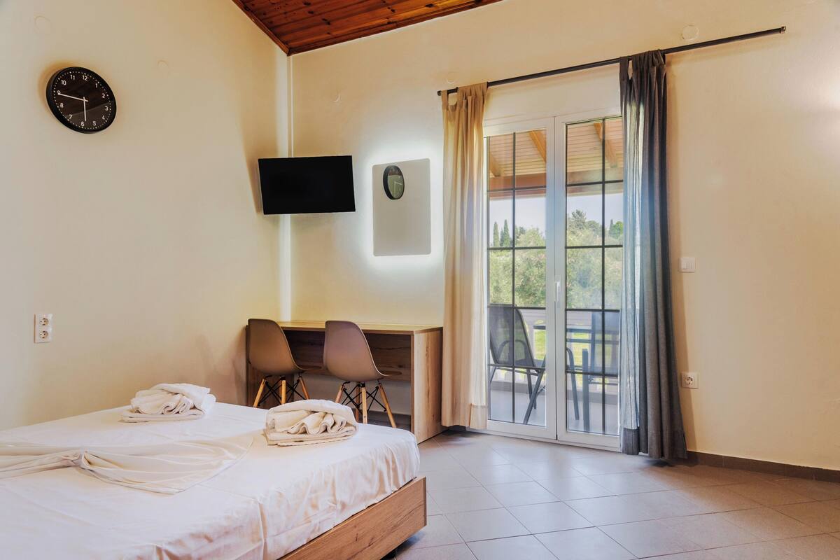 A bedroom with a bed, a desk, two chairs, a TV, a clock, and a sliding glass door to a balcony.
