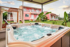 Un jacuzzi con terraza de madera, un área techada con baranda blanca, y un edificio rosa con balcón al fondo.